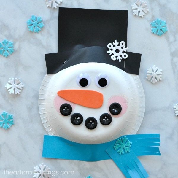 Paper plate with two googly eyes and five buttons forming a smile attached with orange paper as nose and black paper as hat surrounded with blue and white small snowflakes