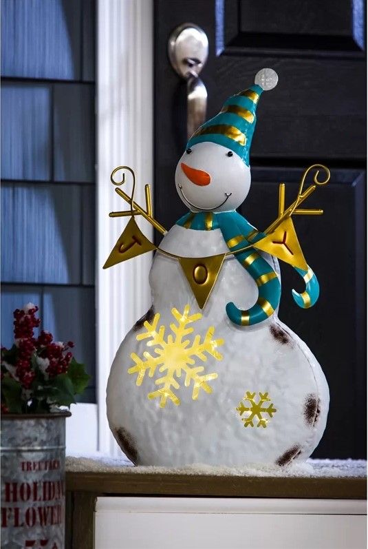 Snowman adorned with gold and blue green decorations holding a bunting with the word JOY in it placed outside