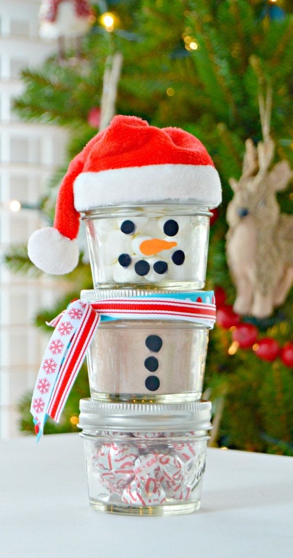 Three jars pile up to form a body of a snowman wearing a santa hat standing on a table with blurred Christmas tree on the background