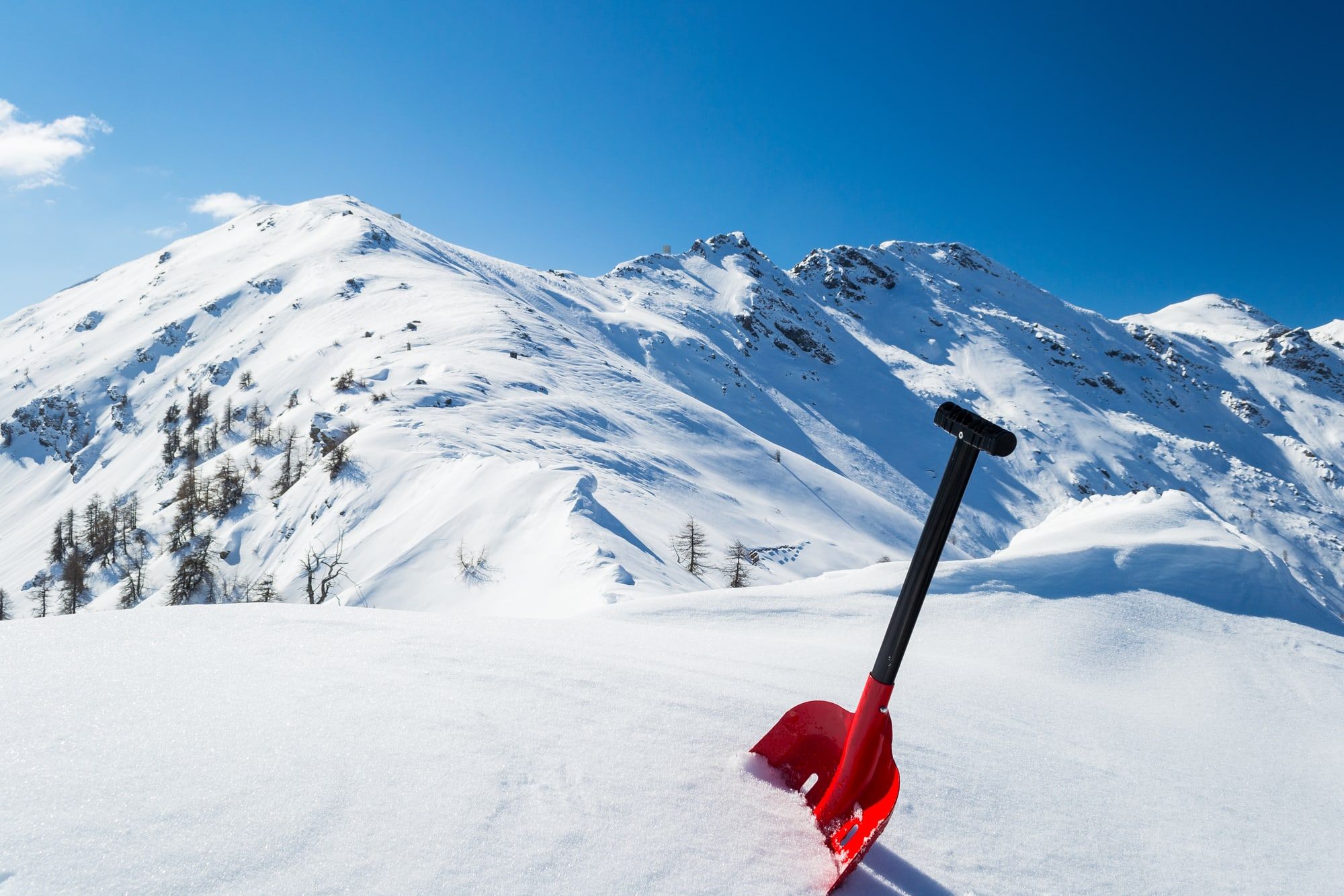 Red avalanche shovel in powder fresh snow. Scenic snowcapped high mountain background. Winter season in the italian Alps.
