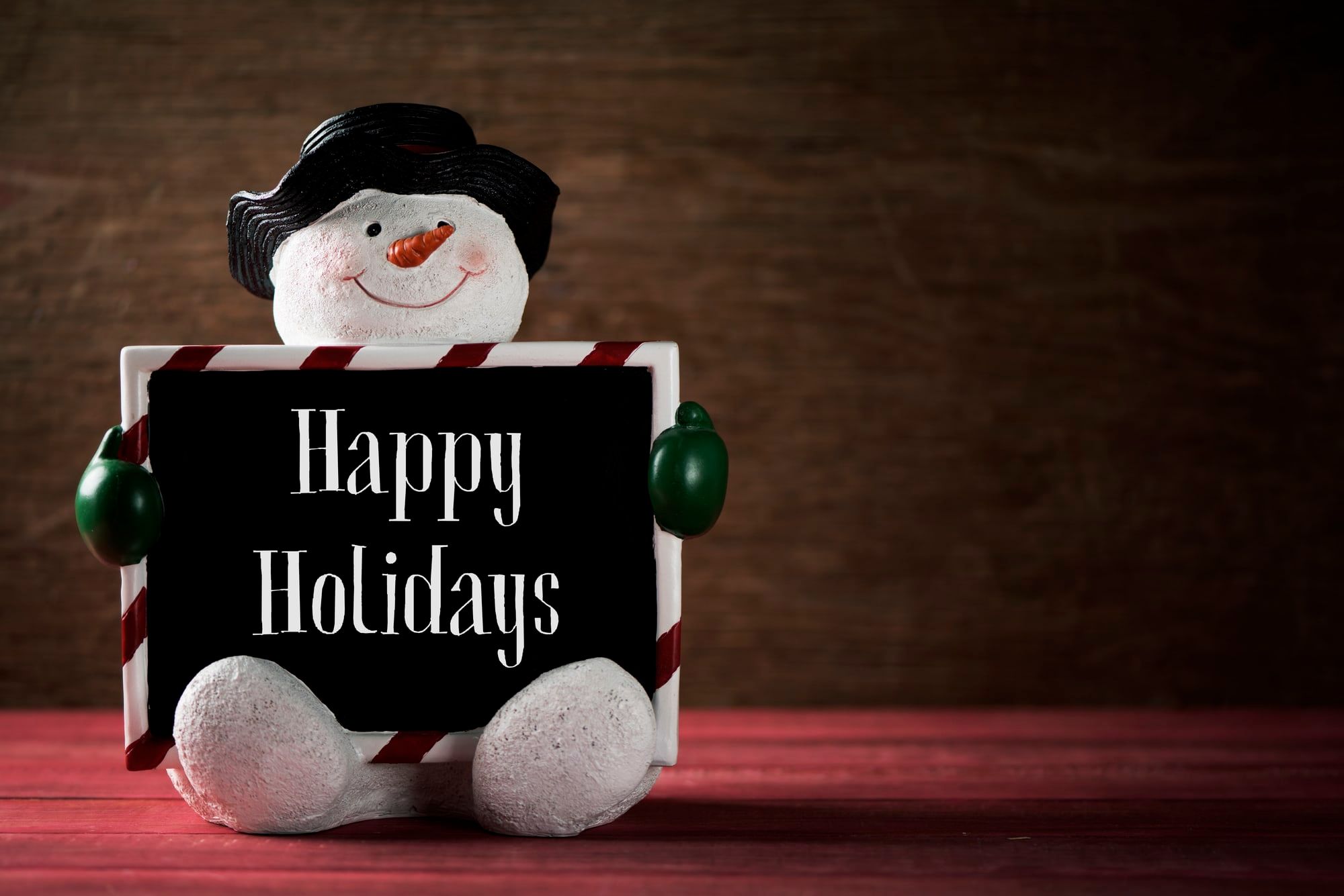 a funny snowman holding a black signboard with the text happy holidays written in it against a rustic wooden surface with a negative space on the right