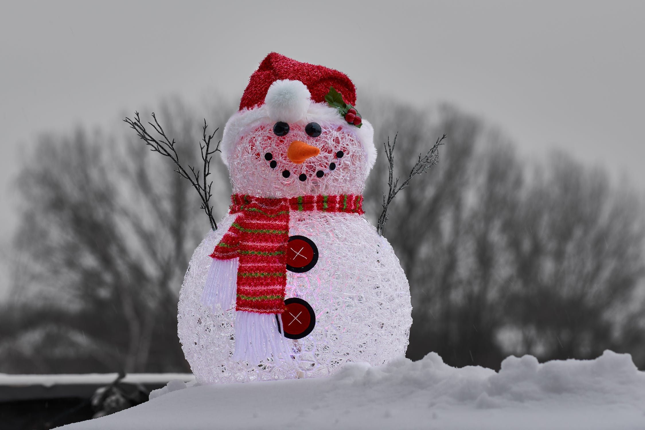 Handmade smiling snowman adorned with red clothings standing on a snow outside with blurred trees on the background