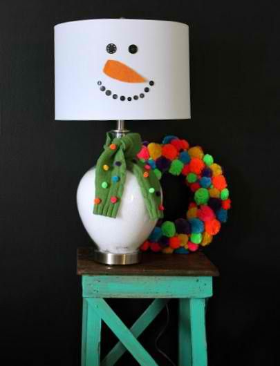A lamp shade with snowman inspired head standing on a small table with colorful pompom on its side