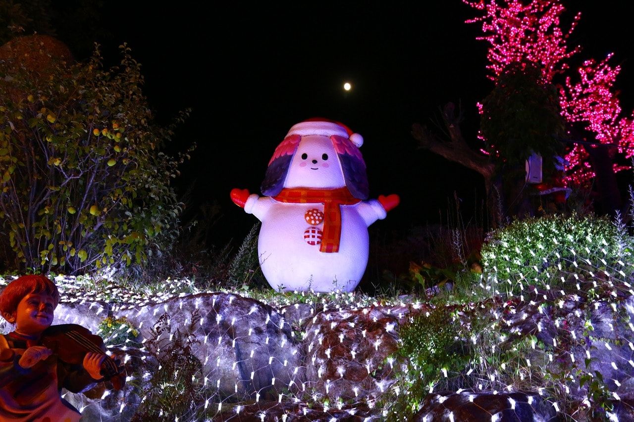 A snowman standing on a christmas light decorated place with trees on its surroundings