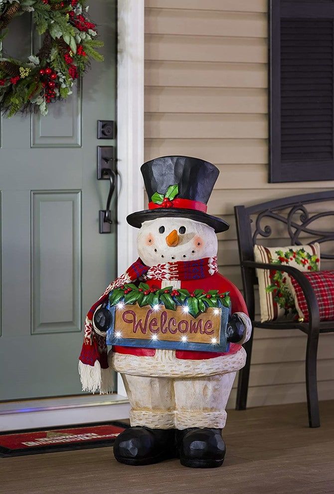 Snowman wearing Santa inspired coat and black hat holding the welcome sign in front of a door and a bench