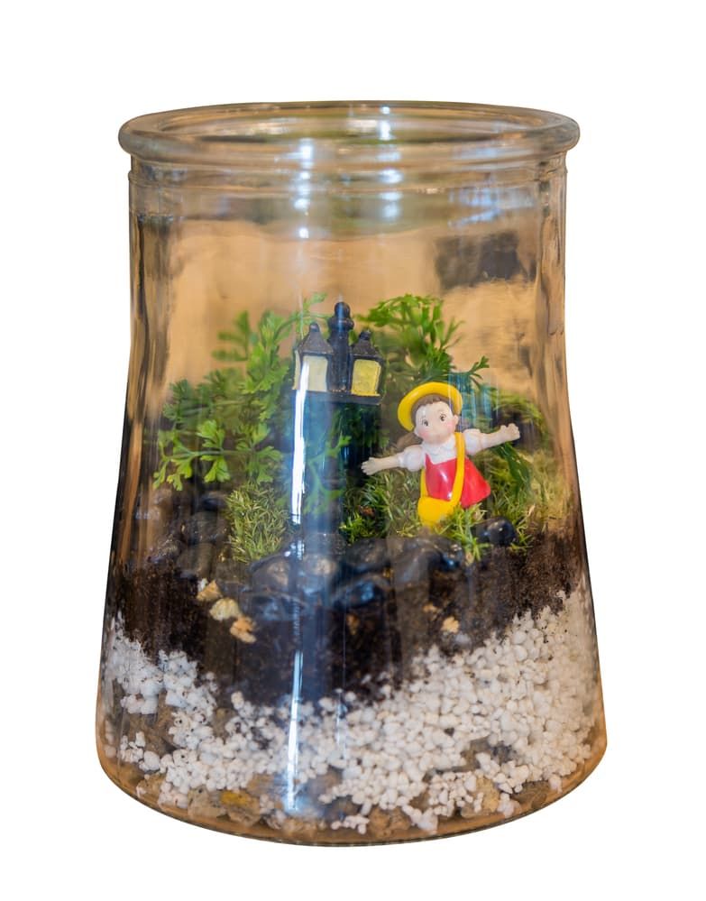 A terrarium with a small garden and a girl doll wearing a pink shirt shoulder bag yellow in a glass jar isolated on white background.