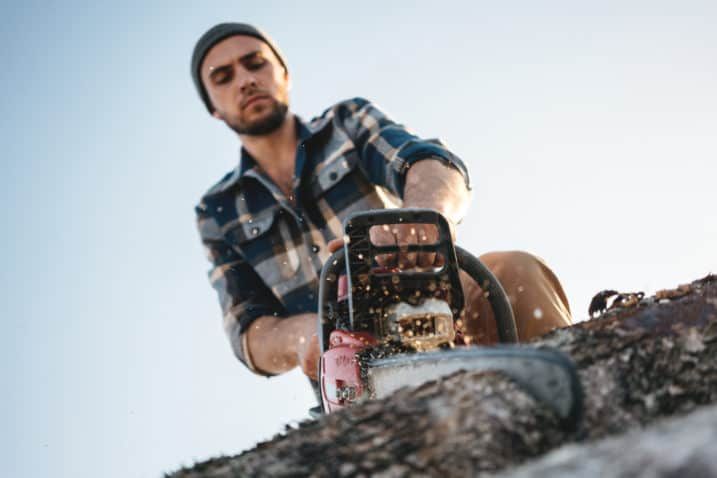 Bearded brutal lumberjack wearing plaid shirt sawing tree with chainsaw for work on sawmill. Wooden sawdust fly apart