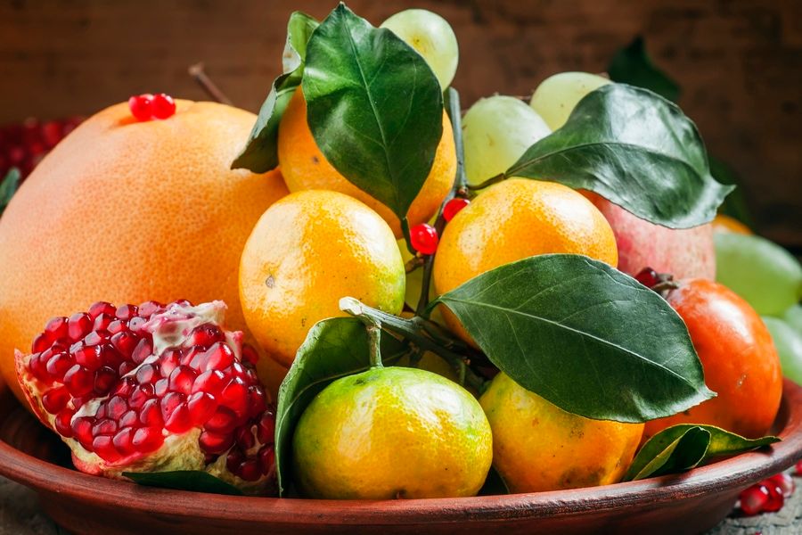 Clay dish with winter fruits: grapefruit, tangerines, persimmons, pomegranates with leaves on the old wooden table, still life, selective focus