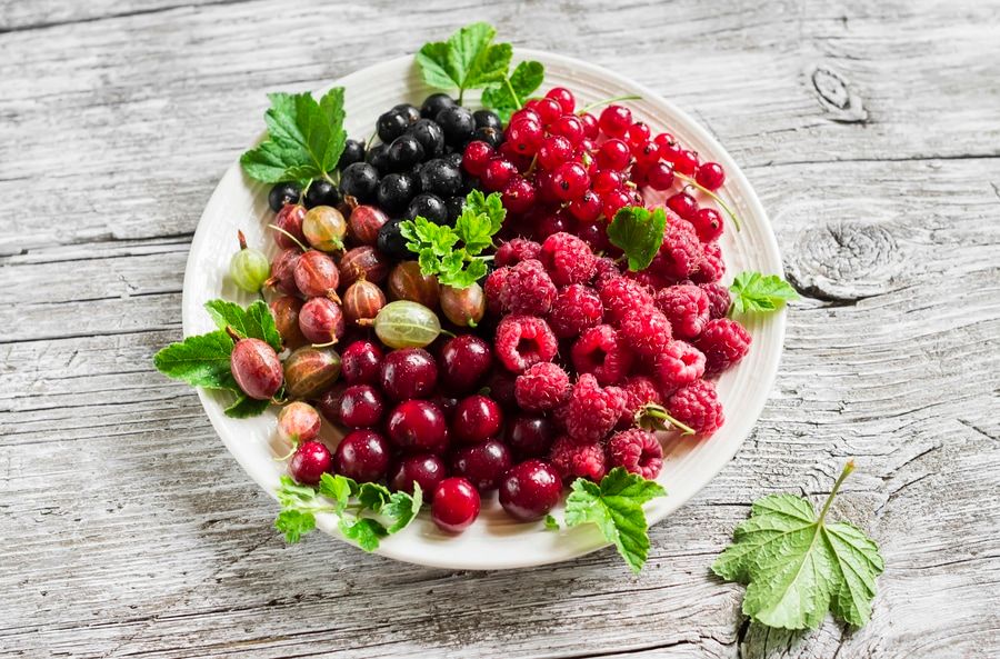 berries - raspberries, gooseberries, red currants, cherries, black currants on a white plate on a light wooden background