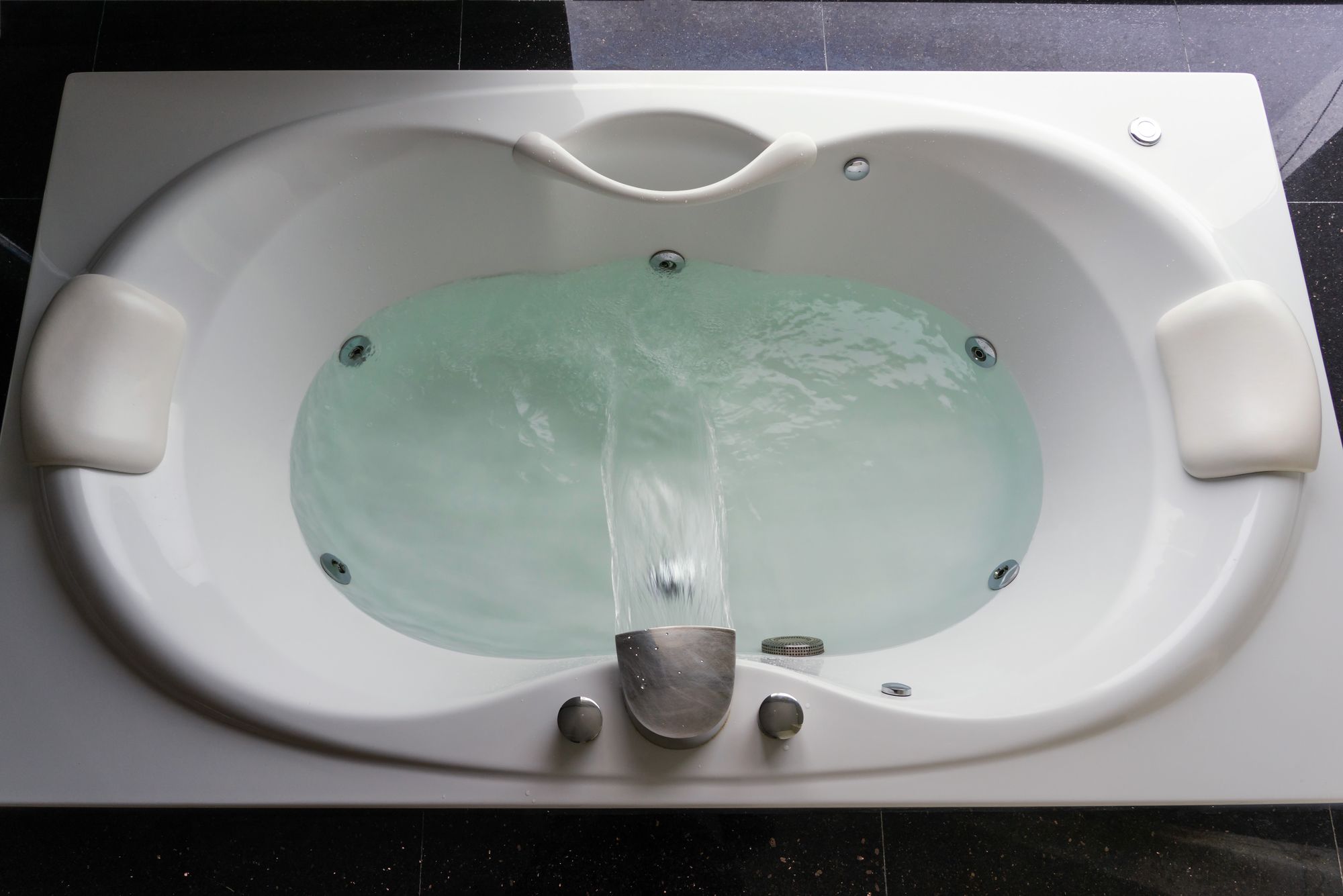 Empty white massaging jetted bathtub with turquoise water on black polished stone floor.