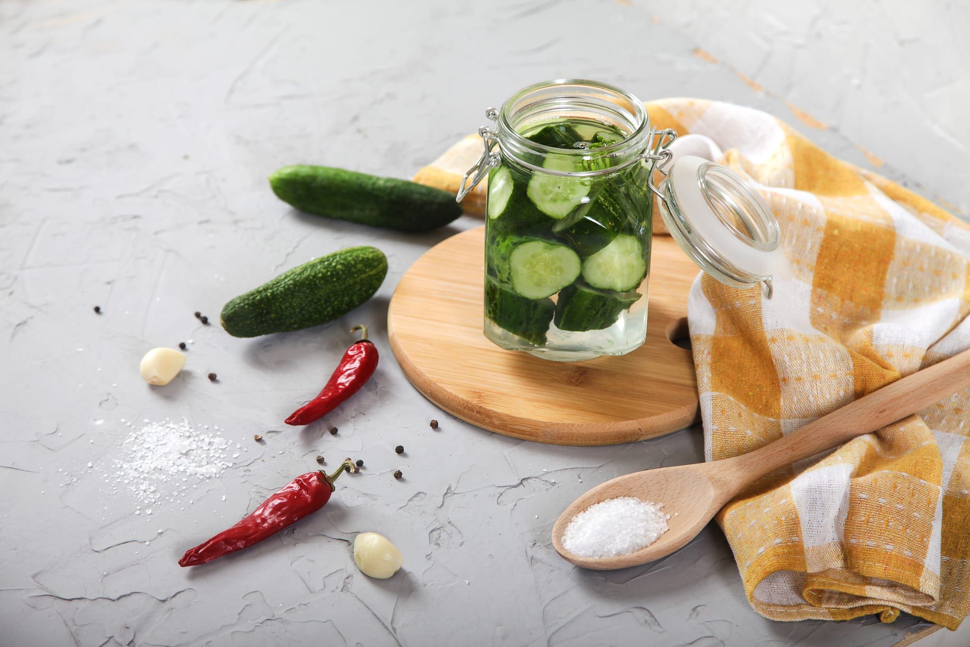 Sliced fermented cucumbers in a glass jar stand on a wooden board. Red hot pepper, garlic, salt in a wooden spoon and a yellow tablecloth on a light gray background