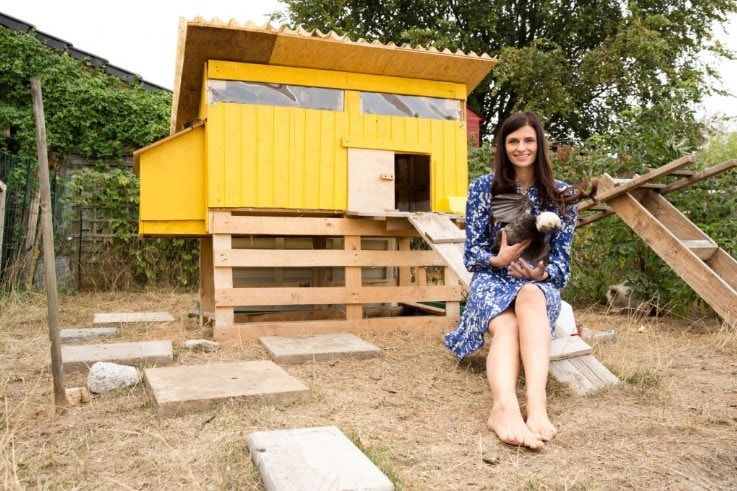 a smiling woman holding a chicken with a yellow chicken coop at her back