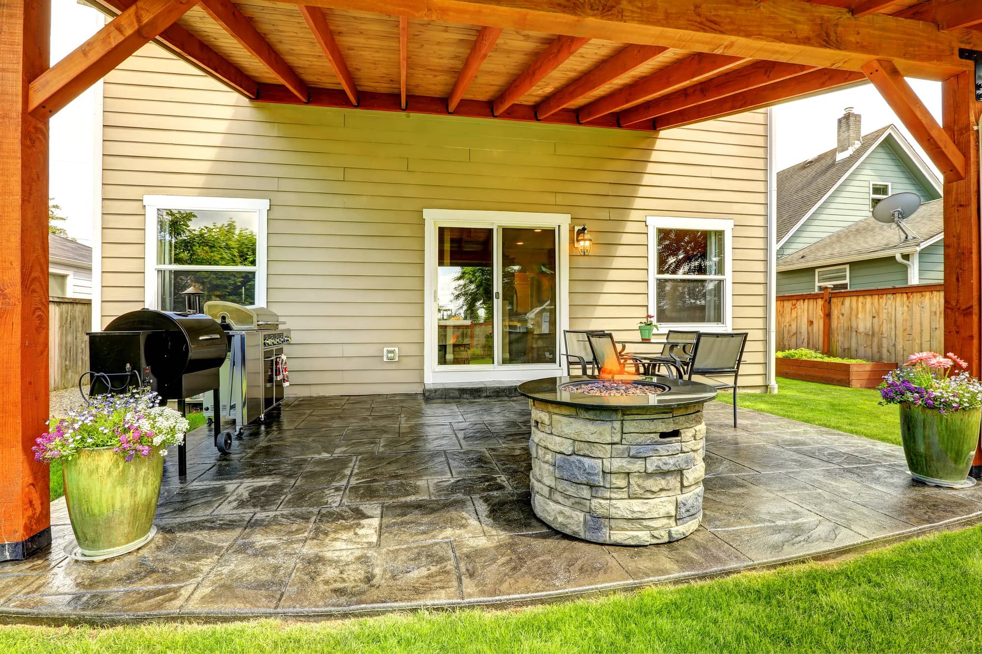 Pergola with patio area. Tile floor decorated with flower pots. Stone trimmed fire pit, patio table set and barbecue