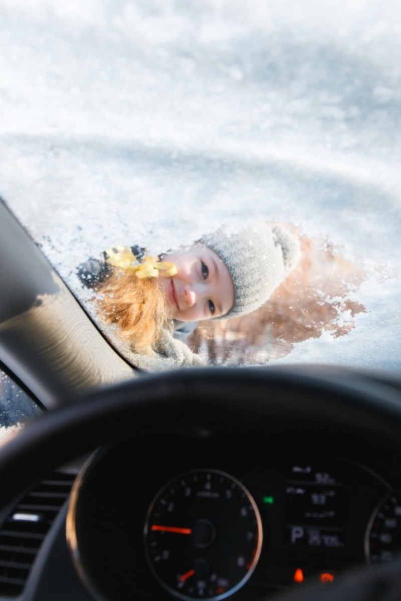 child smiling through a snowy windshield ice scraper clear windows vehicle