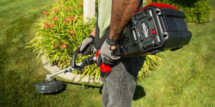 Man holding a battery-powered string trimmer while cutting lawn around a flower bed.