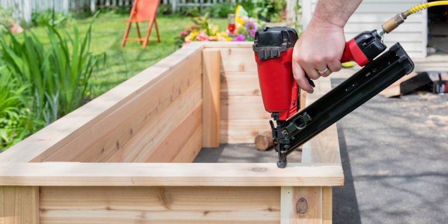 Man using a red nail gun to build a wooden garden bed.
