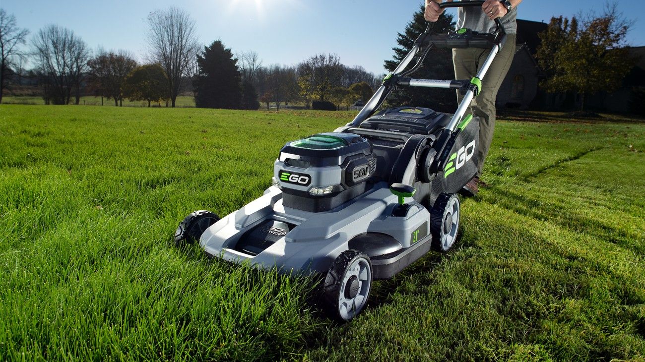 A silver and grey Ego lawn mower with a few vivd green highlights on the motor, levers, and hopper cutting tall grass in a big yard.