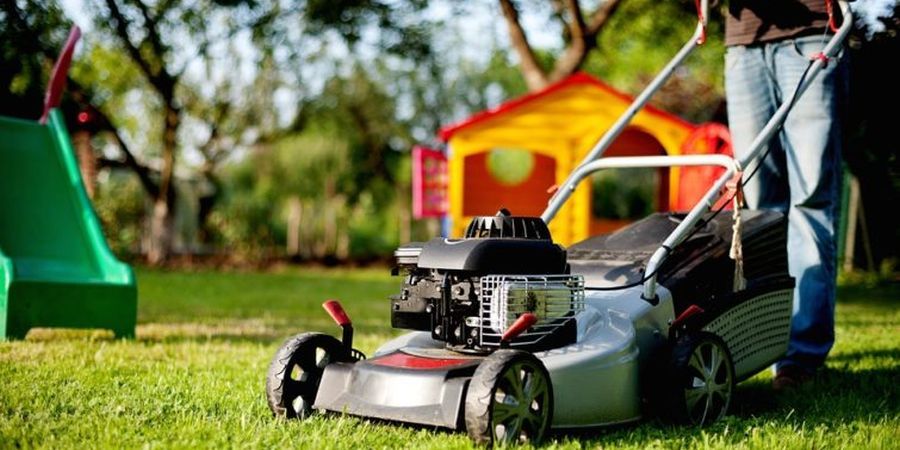 A shiny silver self-propelled lawn mower sitting on the lawn in front of some outdoor children's playground equipment.
