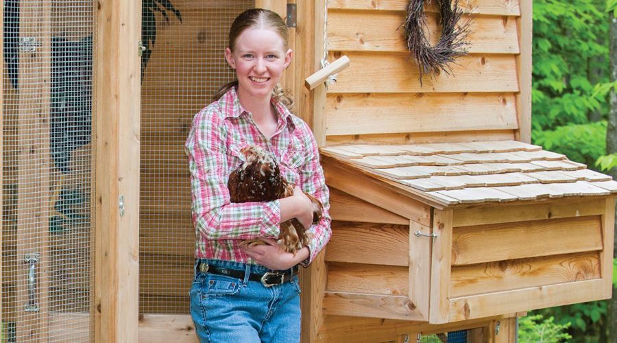 Woman holding a chicken standing outside a large wooden chicken coop.