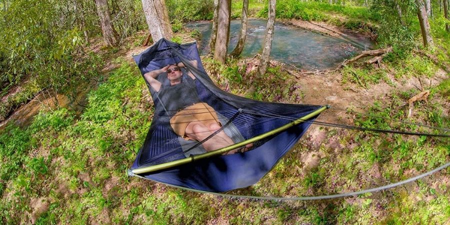 A man wearing black sunglasses, a navy blue t-shirt and brown shorts is laying in a dark blue camping hammock tent with black mosquito netting in a wooded area with a small body of water behind him.