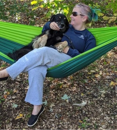 A woman with pink and teal hair in a pony tail, wearing a navy blue long-sleeve t-shirt, grey sweatpants and black shoes is sitting in a green parachute hammock with a black and brown dog next to her and fall leaves all over the ground.