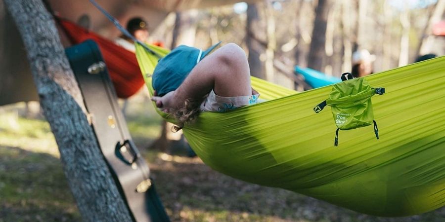 A person stretched out in a lime green parachute hammock with a guitar case leaning against a tree beside them and two other people in an orange hammock and blue hammock in the distance.