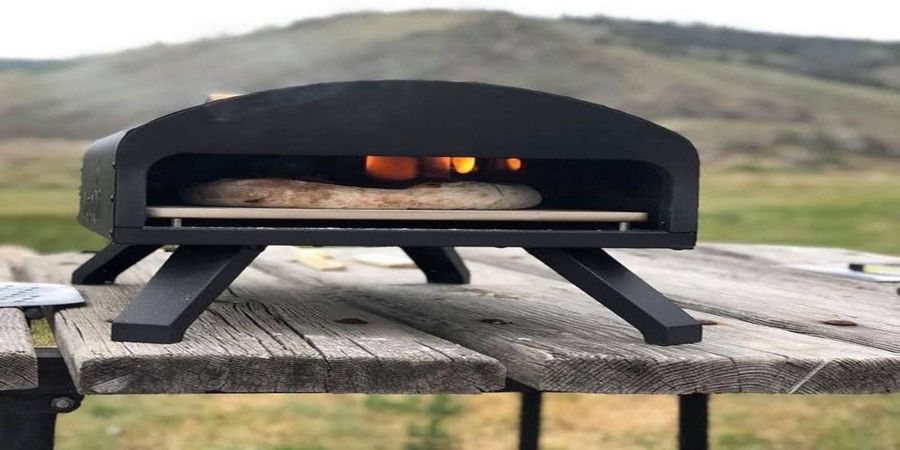 A short black heavy-duty looking outdoor pizza oven sitting on a picnick table with mountains in the background.