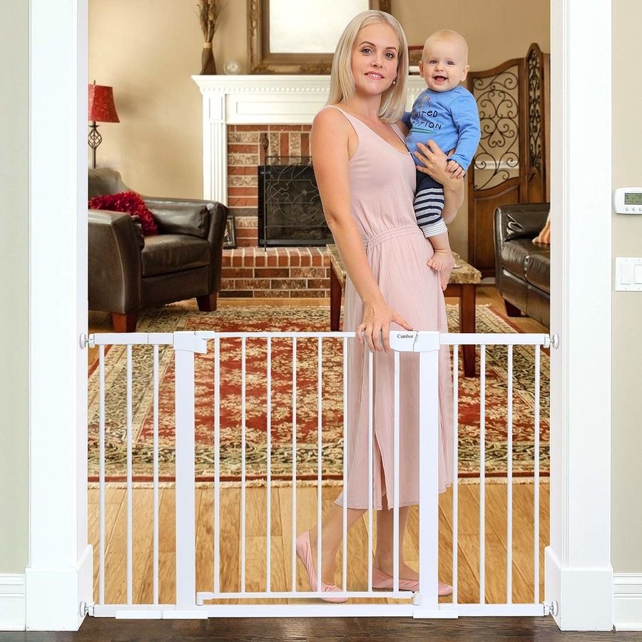 A mother with toddler in one arm stands with her other hand on a wide white dog gates top latch.