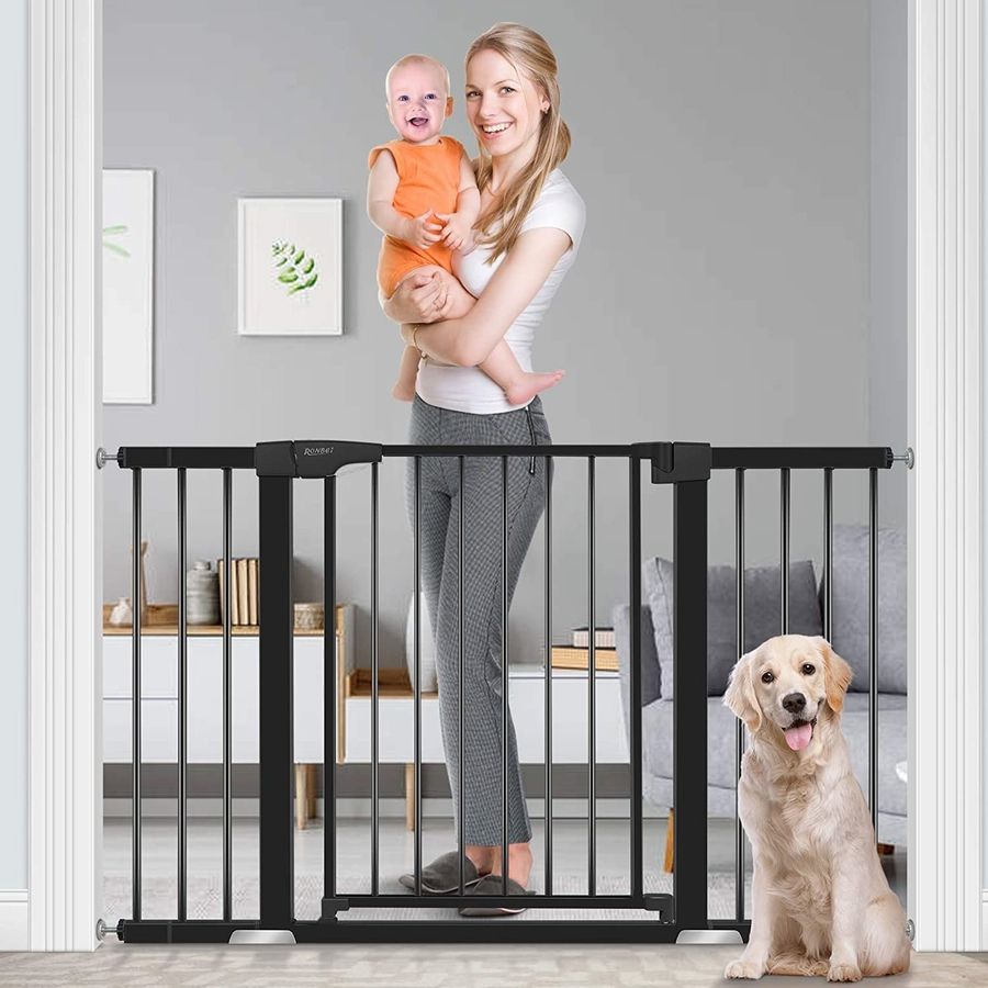 A woman with red hair, with baby in hand, stand on one side of an extra-wide black dog gate, and a blonde dog sits calmly on the other side.
