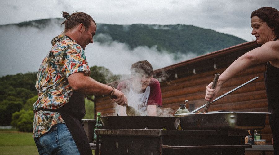 Three adults cooking around an outdoor BBQ with mountains in the background