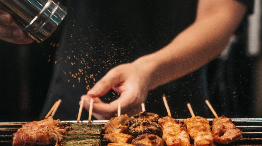 A person shaking spices on shishkabobs on the grill