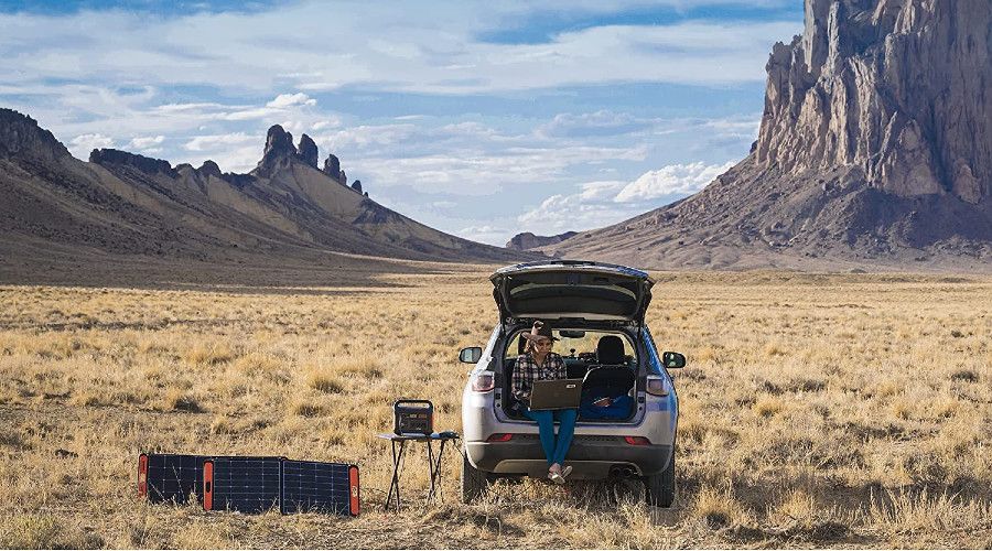woman sitting in back of hatchback car in wilderness, solar energy kit set up in foreground, mountains in background
