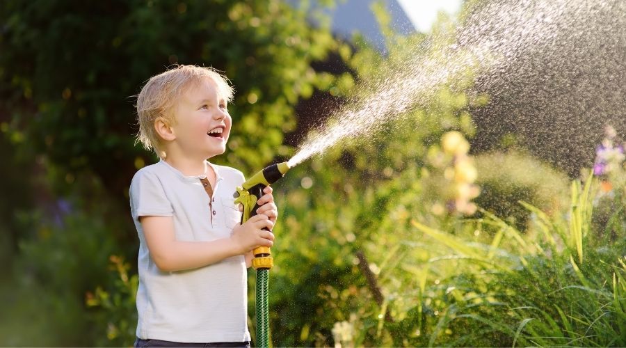 young boy holding the sprayer end of a garden hose, watering the garden
