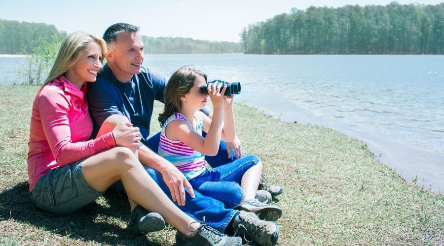 family of three sitting on the shore of a lake with the child looking through binoculars