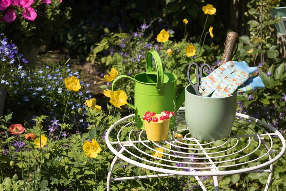 View of colorful flowering summer garden. Garden tools, gloves, flower pots, watering can is on a garden table among growing flowers on a sunny day. Concept: gardening.