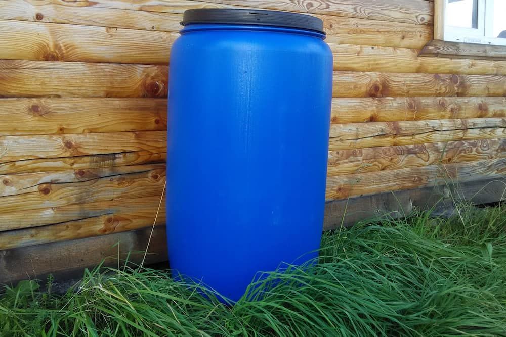 large blue plastic barrel with a black lid stands on the green grass against the wall of a log house