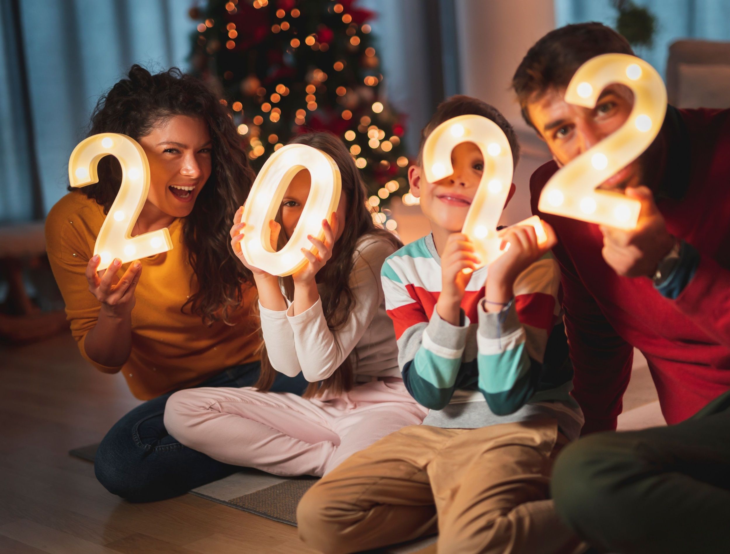 Happy family celebrating New Years Eve at home with kids, sitting by the Christmas tree, holding illuminative numbers 2022 representing the upcoming New Year