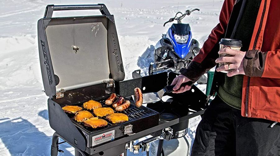 Man cooking meat on a Camp Chef BB30L grill in a winter wonderland with snowmobile in the background.