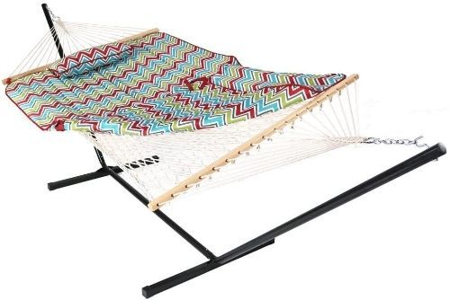 A white rope style hammock hanging from a black metal hammock stand. On the hammock is a green, blue and pink chevron hammock pad and pillow.